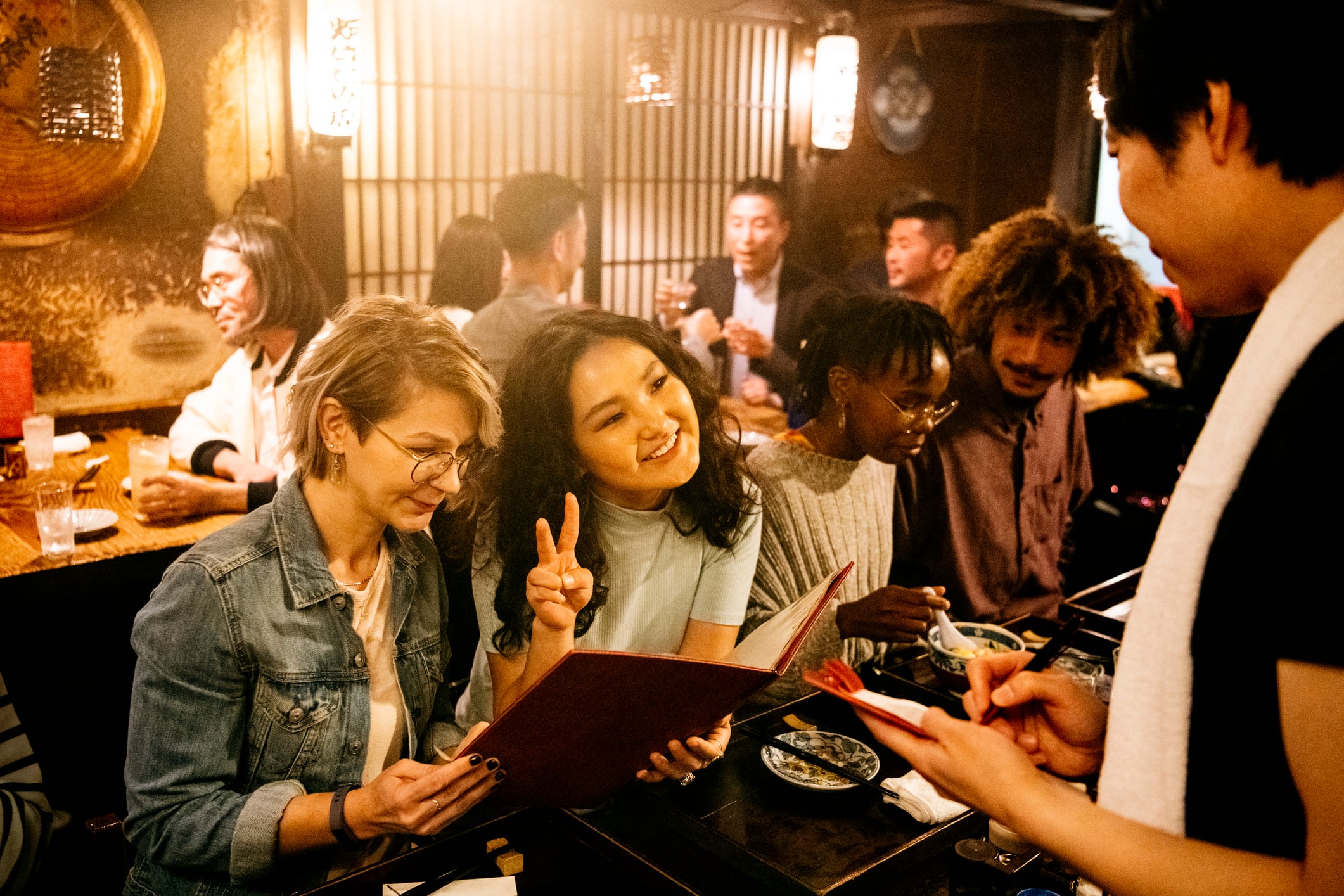 Waiter in Izakaya taking food order from attractive young women choosing from menu, making peace sign, dining, fun, leisure