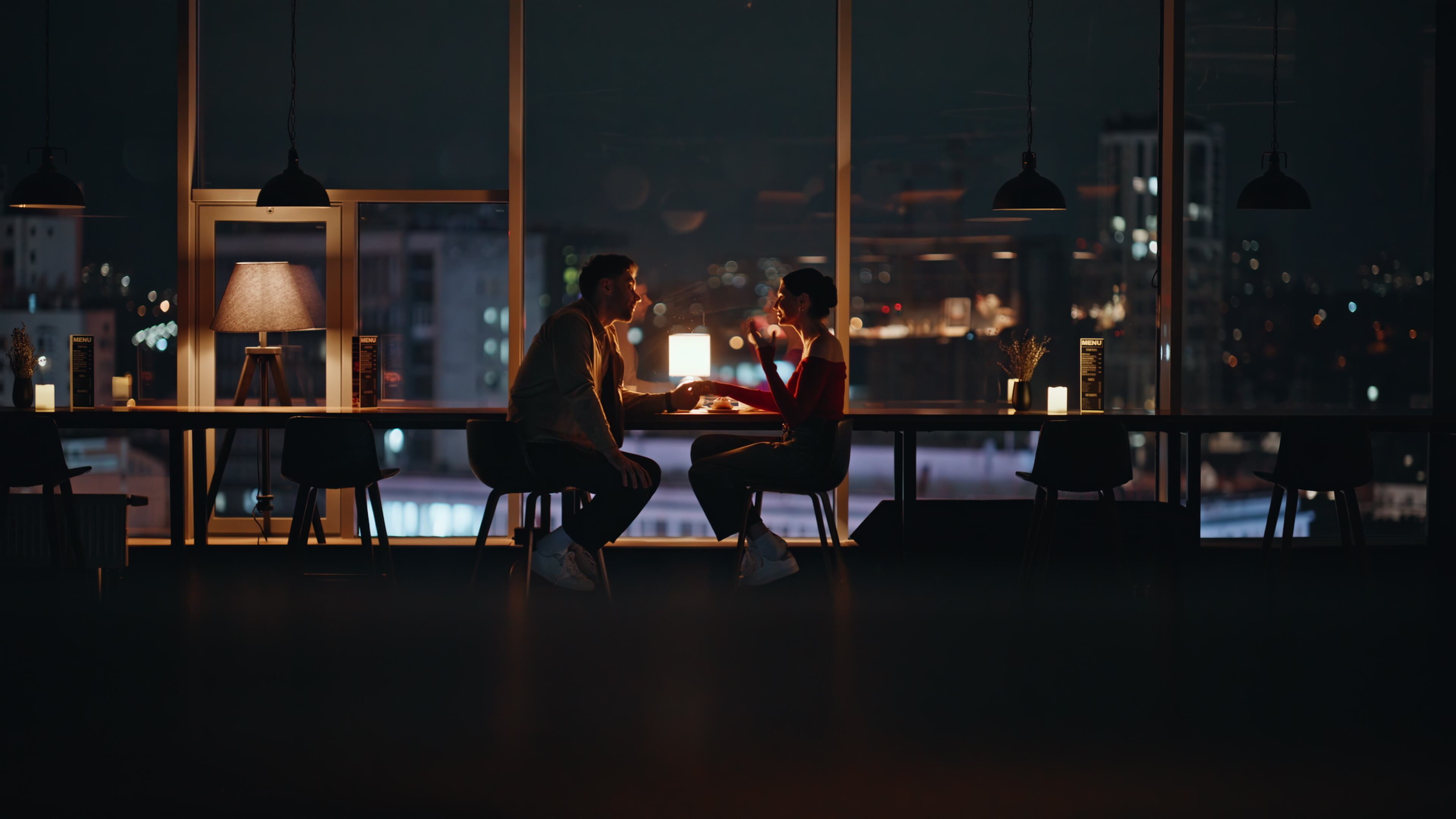 Couple dining in cozy restaurant with night city view. Happy spouses talking at romantic evening date sitting luxury cafe table together. Love pair enjoy relationship spending time in dark cafeteria.